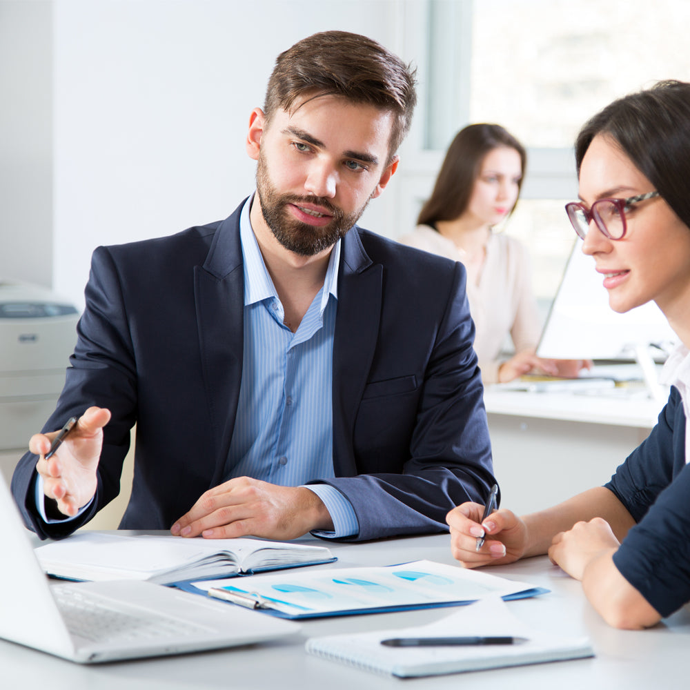 Man in a suit explaining something to a woman at a desk with documents and a laptop.