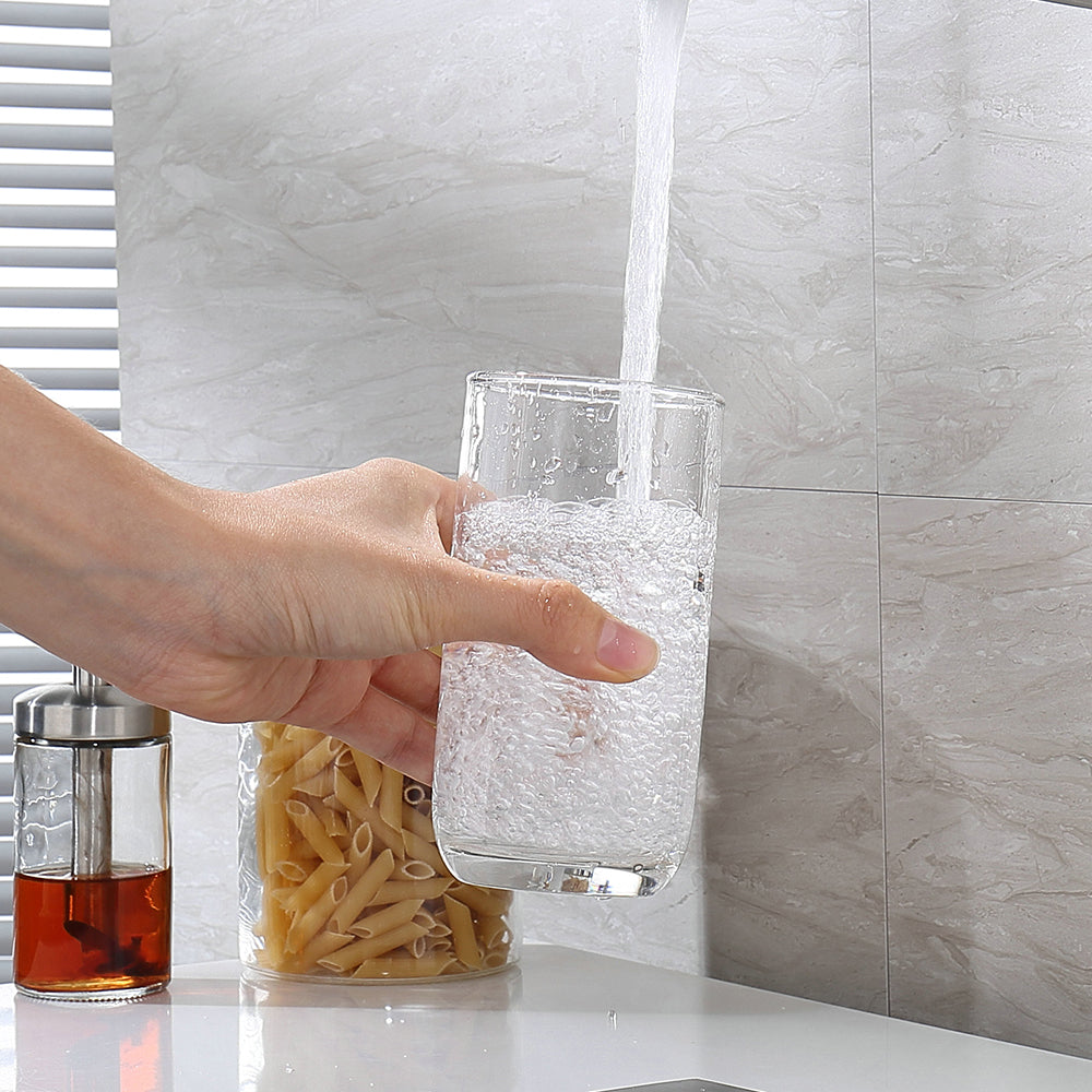 Hand holding a glass under running water with pasta and a tea bag in the background.